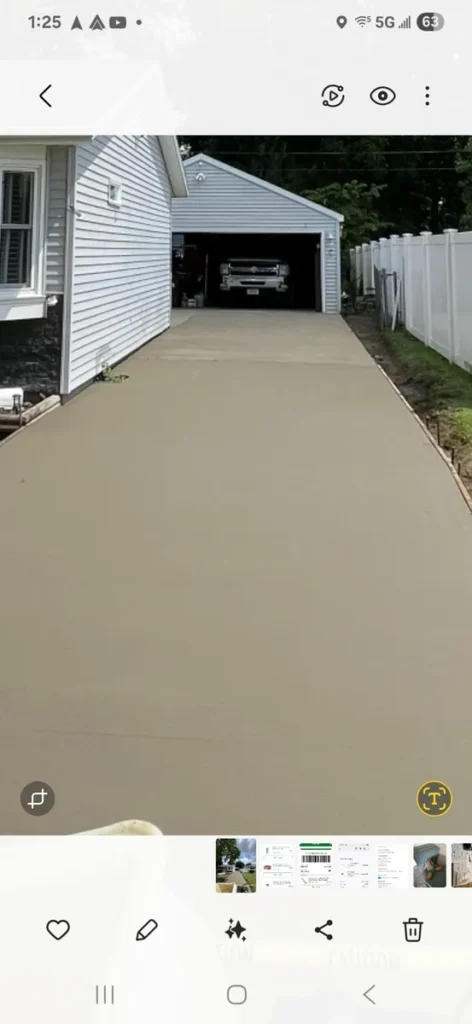 New concrete driveway leading to an attached garage with a truck parked inside, white vinyl fence on the right