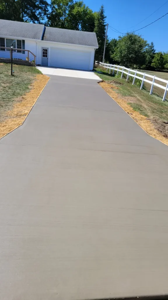 Long smooth concrete driveway leading to a white detached garage beside a ranch-style home with a white wooden fence