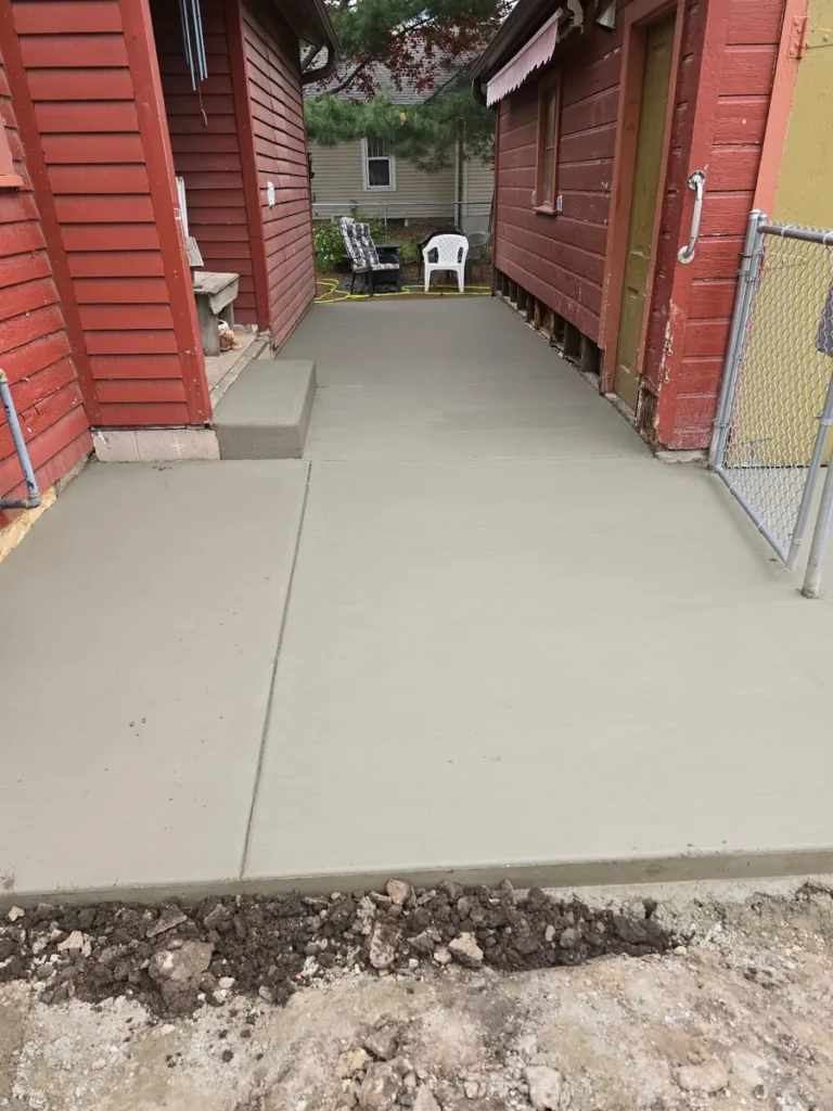 Freshly poured concrete walkway and pad between two red wood-sided buildings with a chain-link gate