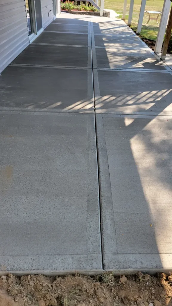 New brushed-finish concrete sidewalk with control joints running alongside a house, shaded by a porch