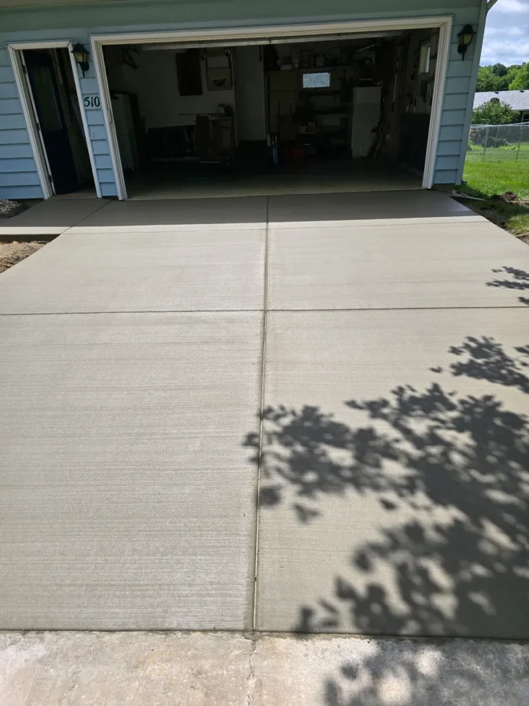 Fresh concrete driveway apron in front of a blue two-car garage with tree shadows across the surface