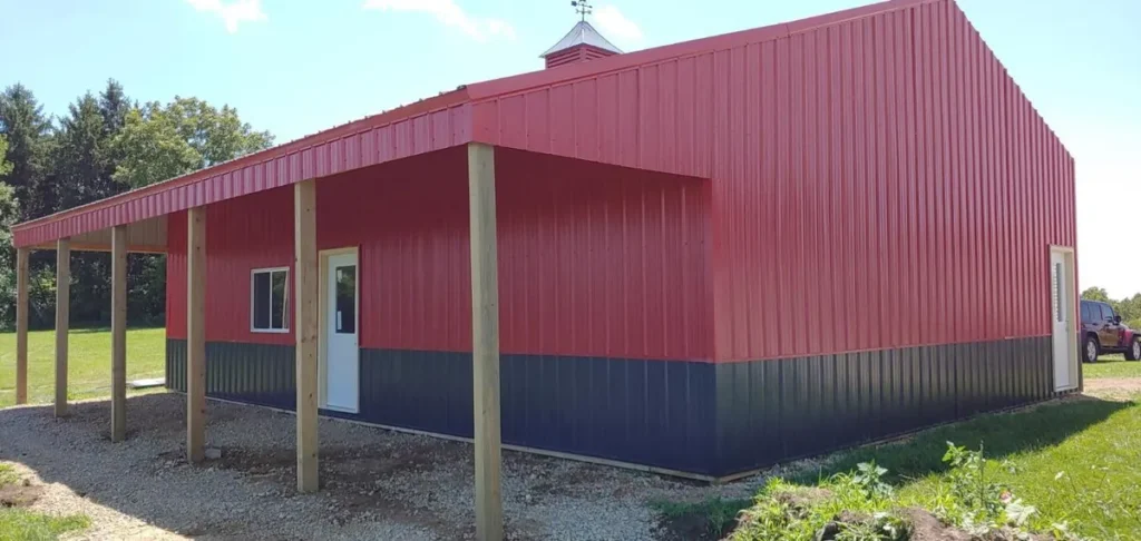 Red and dark blue metal pole barn with a lean-to overhang supported by wood posts on a gravel base