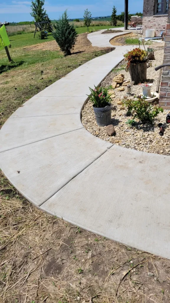 Curved concrete sidewalk with expansion joints winding past a flower garden and rock border along a brick house