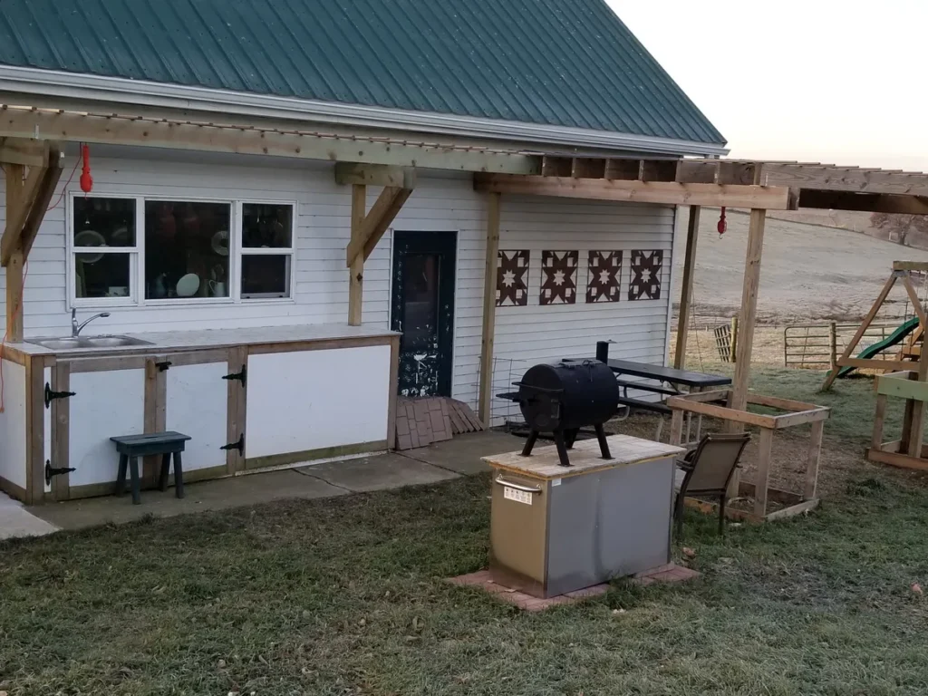 Outdoor kitchen setup with a concrete pad, sink counter, smoker grill, and pergola frame attached to a white building