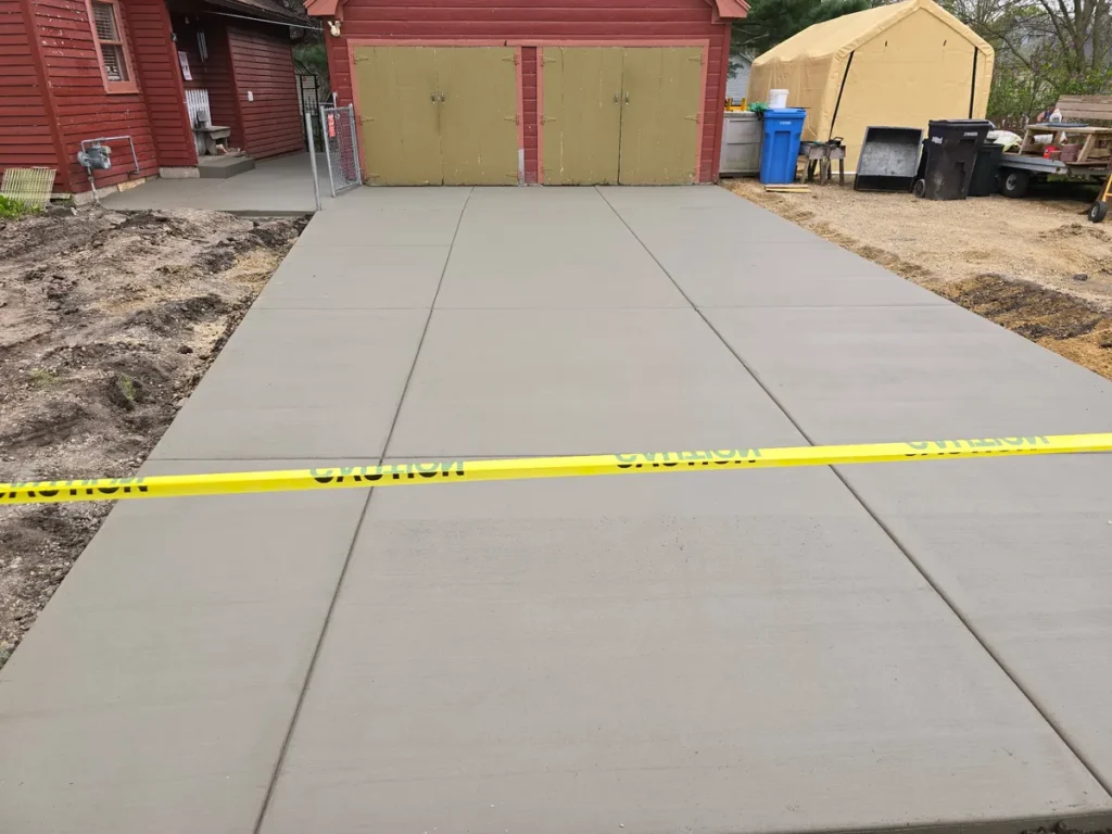 Freshly poured concrete driveway in front of a red building with yellow caution tape marking the fresh concrete edges