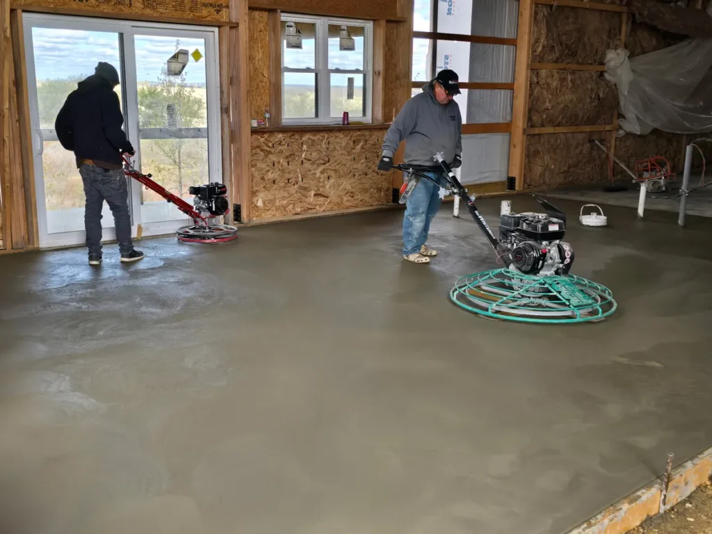 Two workers operating power trowel machines to finish a concrete floor inside a wood-framed building under construction