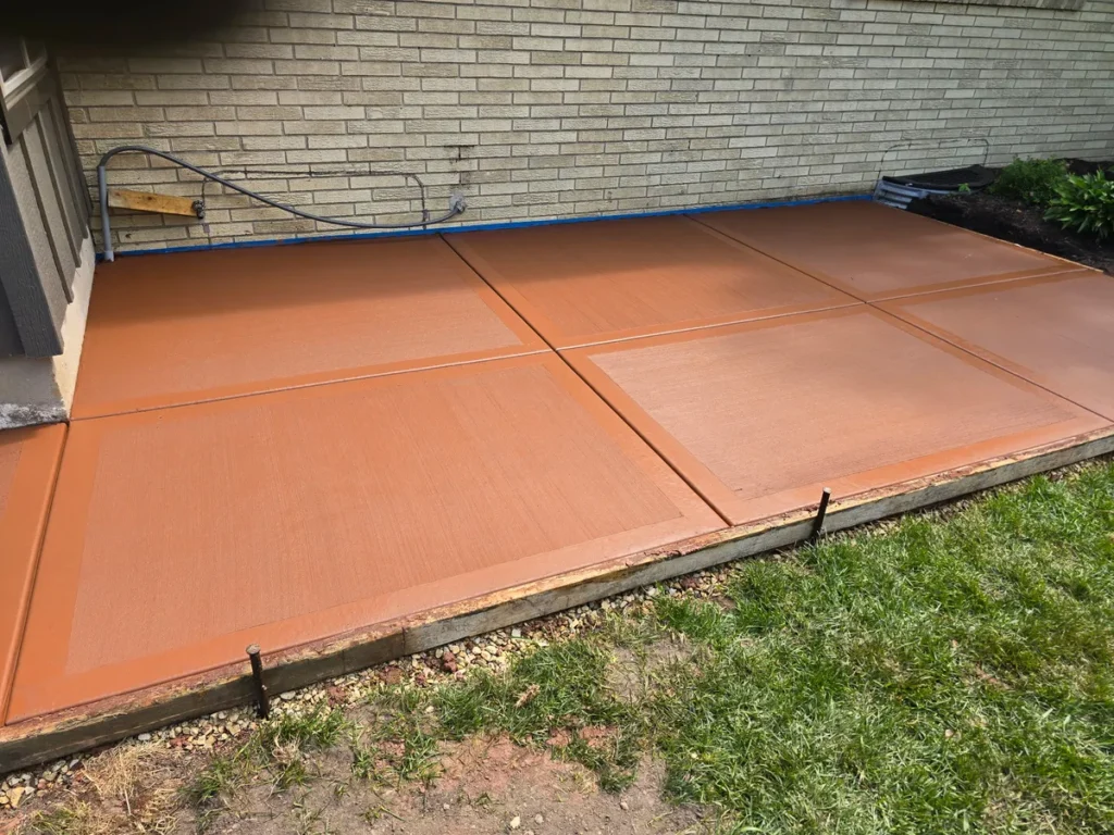 Concrete patio with terracotta red acid stain and control joints beside a brick house