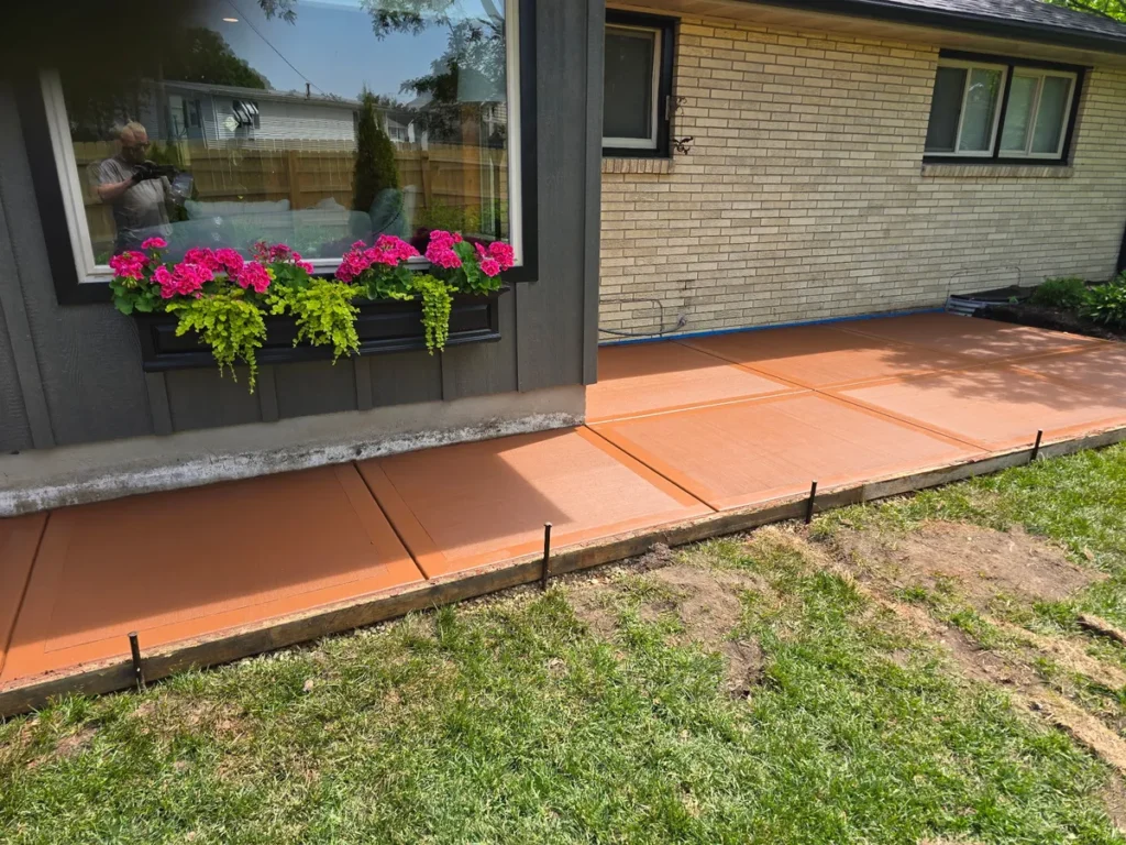 Terracotta red acid-stained concrete patio with control joints beside a brick house, flower box with red geraniums visible