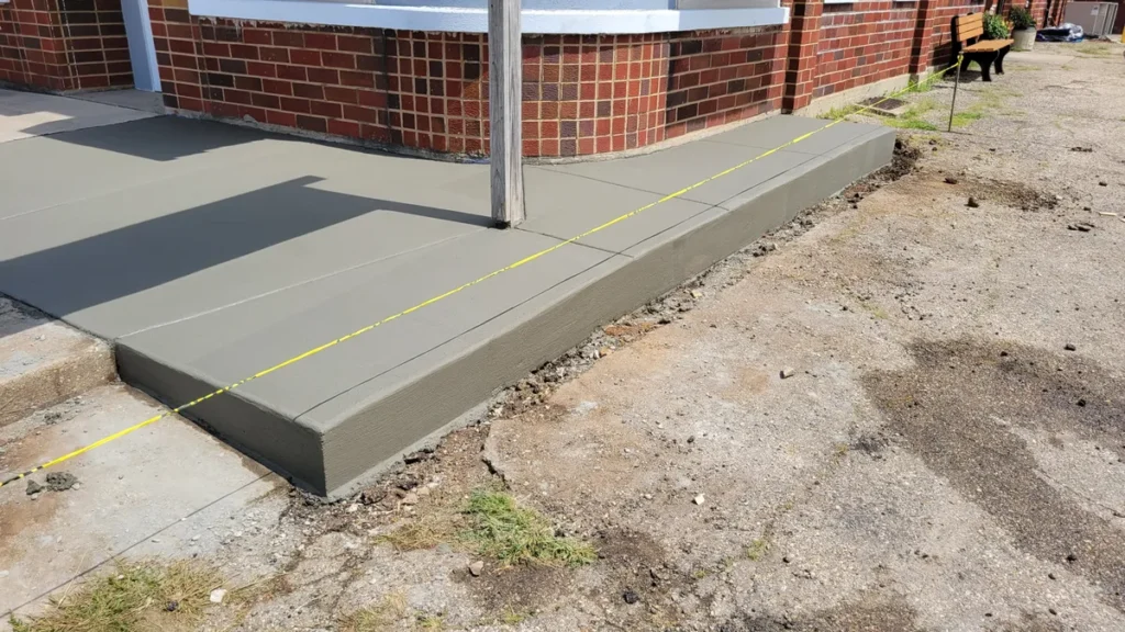Newly poured concrete ramp addition at a brick building entrance, marked with yellow caution tape