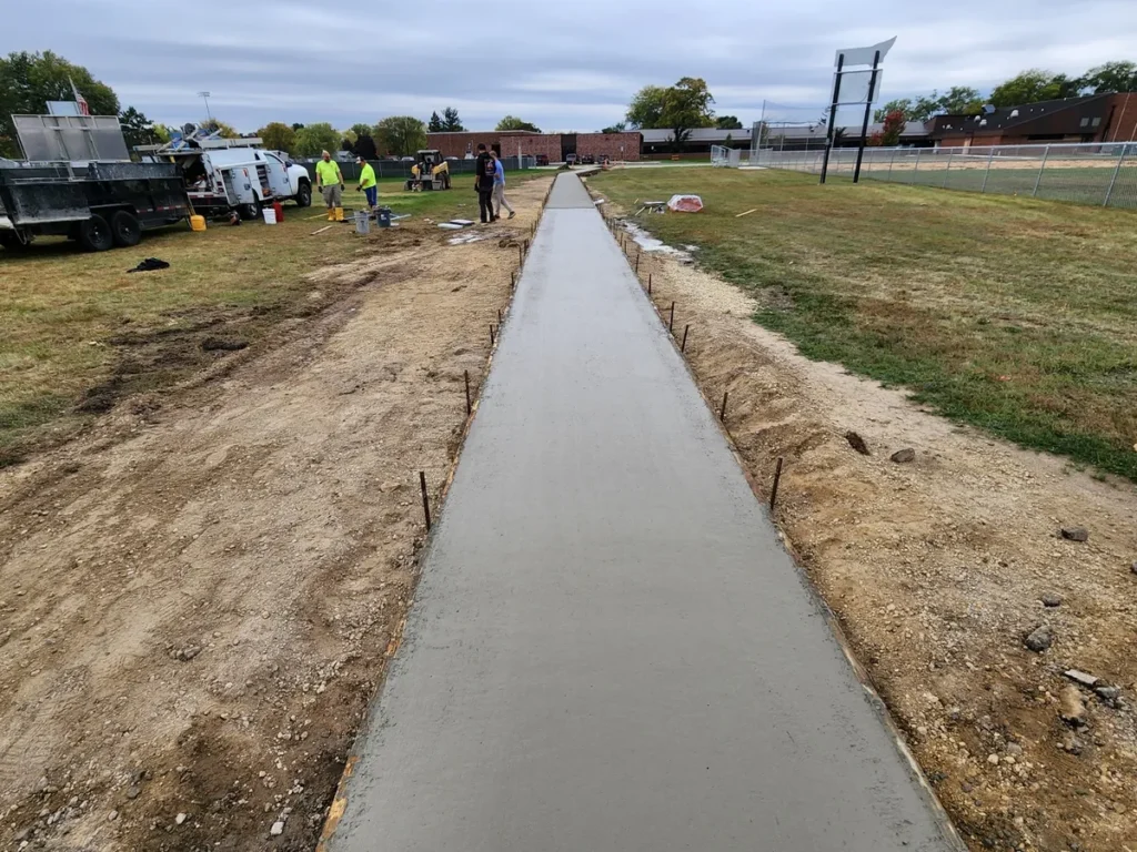 Construction crew pouring a long straight concrete sidewalk through a school athletic field, concrete trucks and workers visible