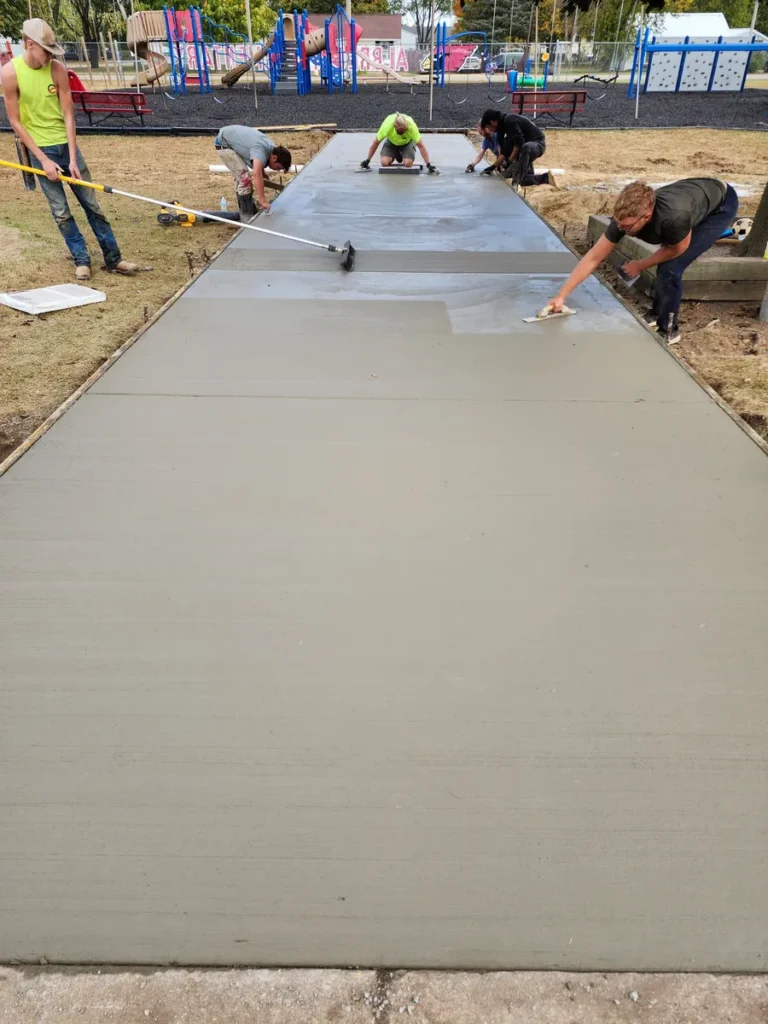 Crew of workers hand-finishing a freshly poured wide concrete sidewalk pad near a school playground