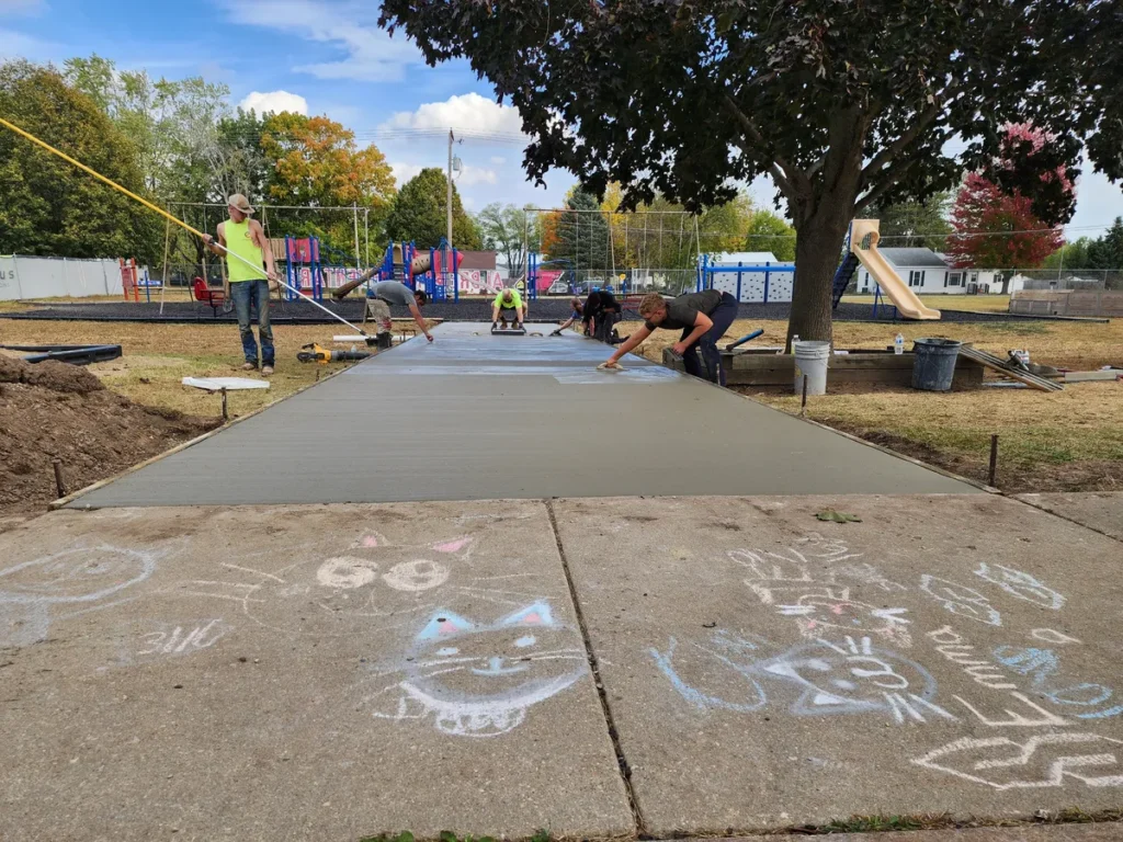 Crew finishing a fresh concrete sidewalk near a playground, chalk drawings visible on the adjacent existing concrete