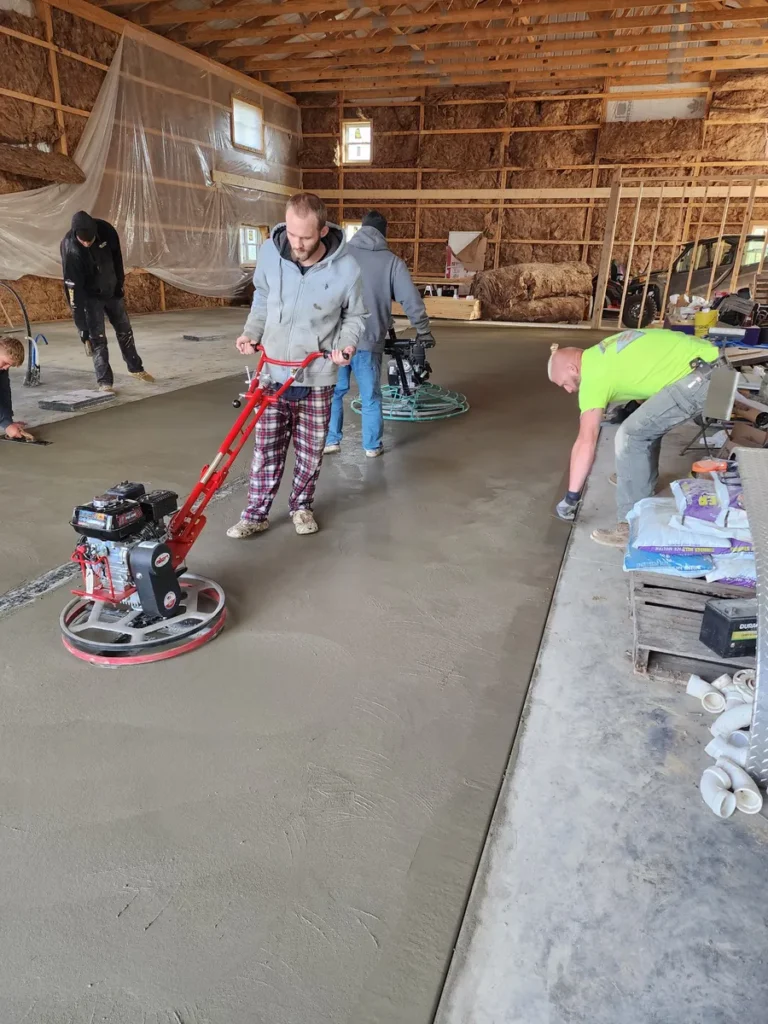 Workers using a power trowel and hand tools to finish a freshly poured concrete floor inside a barn with insulation on the walls