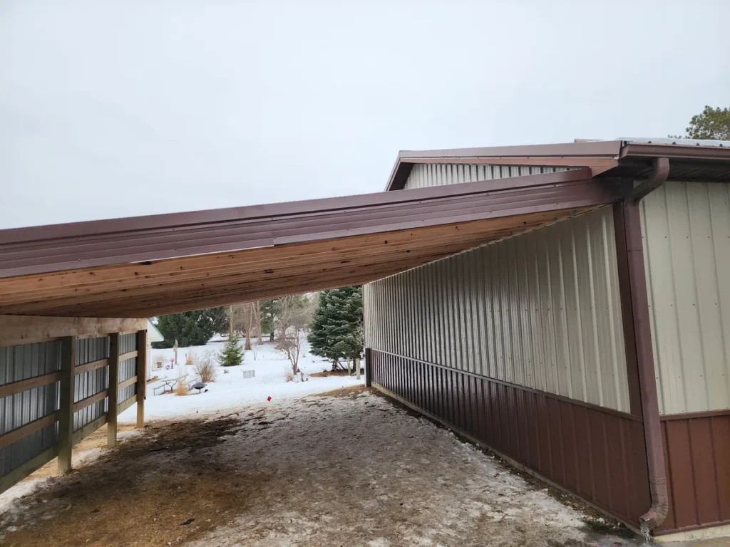 Completed lean-to carport with cedar log rafters and brown metal roof attached to a tan and brown metal pole building, snow on ground